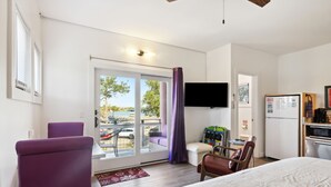 A hotel room with a bed, a purple armchair, a wooden chair, a small table, a flat-screen TV, and a view of the parking lot through the sliding glass door.