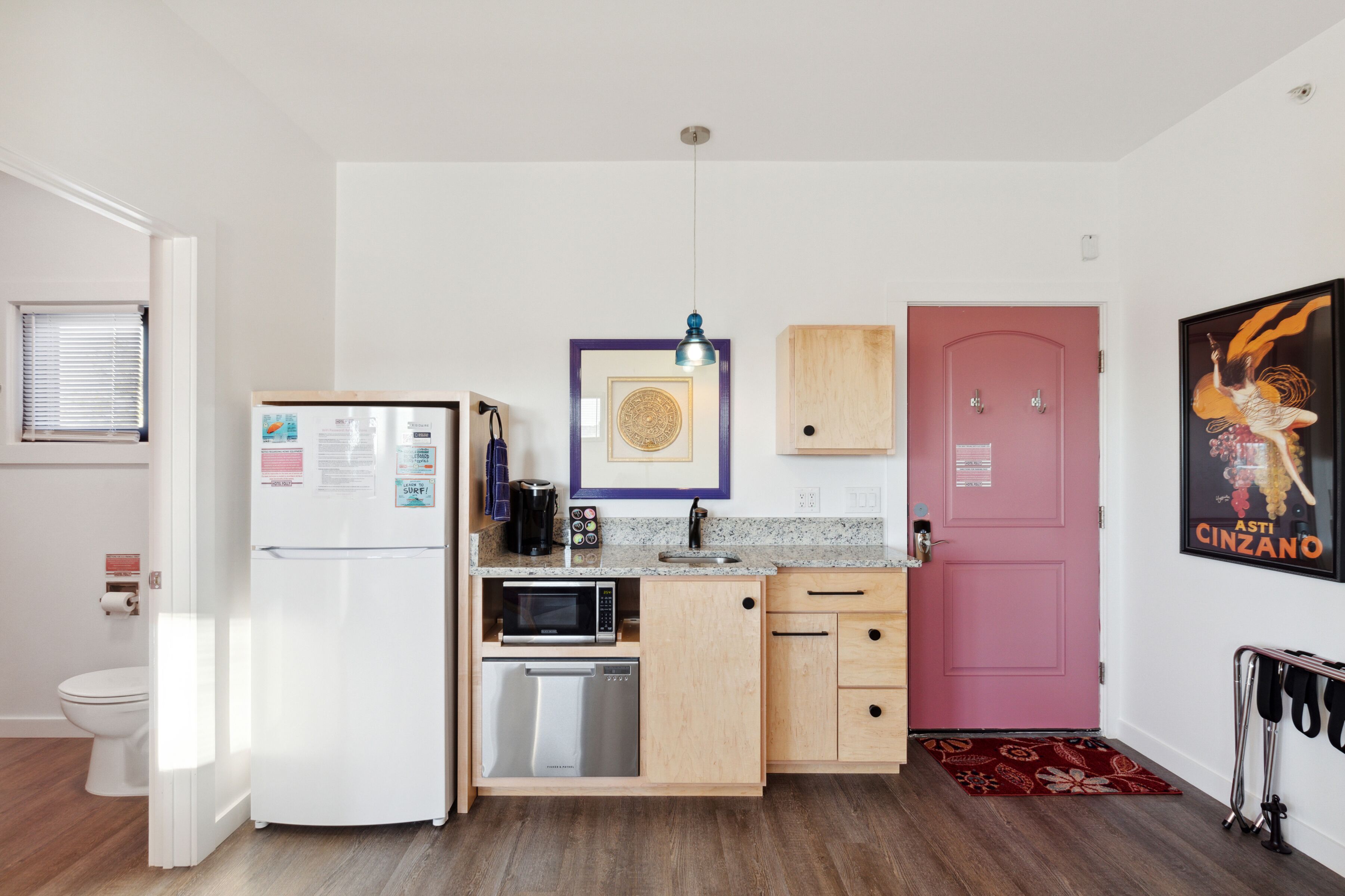 A compact kitchen with a white refrigerator, microwave, and dishwasher, wooden cabinets, a pink door, and a poster on the wall.