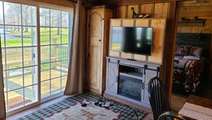 A rustic cabin interior with a large window, wooden walls, a fireplace, and a mounted television.