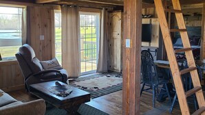 A rustic cabin interior with a wooden ceiling, a ceiling fan, a large window, a cozy seating area with a chair and a table, and a wooden ladder.