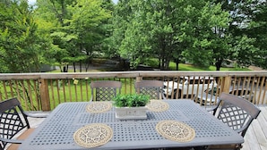 Une terrasse avec une table et des chaises, entourée d’une rambarde en bois, donnant sur un jardin avec des arbres et une voiture blanche.