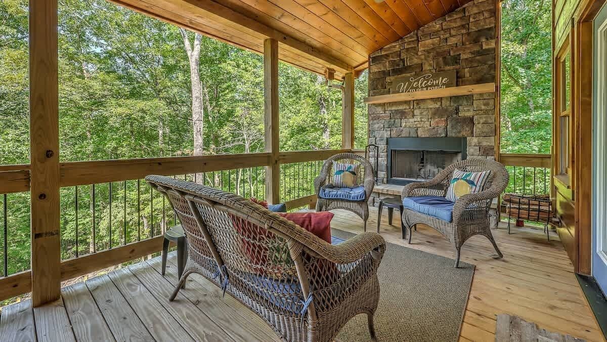 A covered porch with wicker furniture, a stone fireplace, and a view of the forest.