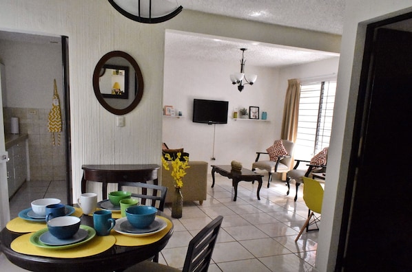 A dining area with a round table set for four, a television mounted on the wall, and a living area with a sofa and chairs.
