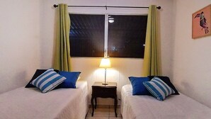 Ein Zimmer mit zwei Einzelbetten, einem Nachttisch mit Lampe, einem Fenster mit Vorhängen und einem Wandbild.