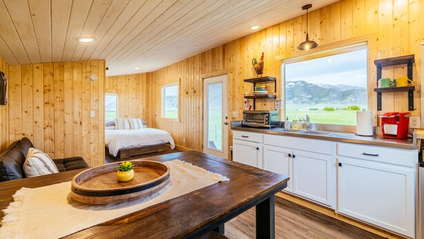 A cozy room with a bed, a wooden table with a tray, a kitchen with white cabinets, and a window with a mountain view.