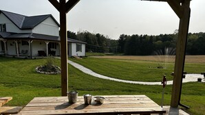 A wooden picnic table with metal buckets and a water spout in a covered outdoor area with a view of a house and a grassy field.