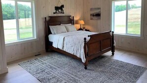 A bedroom with a wooden bed, a patterned rug, and large windows offering a view of the outdoors.