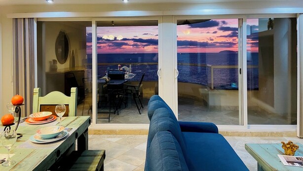 A balcony with a view of the sea at sunset, a dining table with plates and glasses, and a blue sofa.