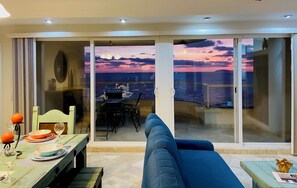 A balcony with a view of the sea at sunset, a dining table with plates and glasses, and a blue sofa.