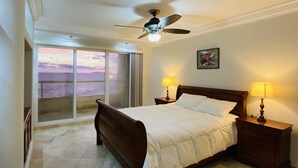 A bedroom with a bed, two bedside tables with lamps, a ceiling fan, a large window with a view of the sea, and a framed picture on the wall.