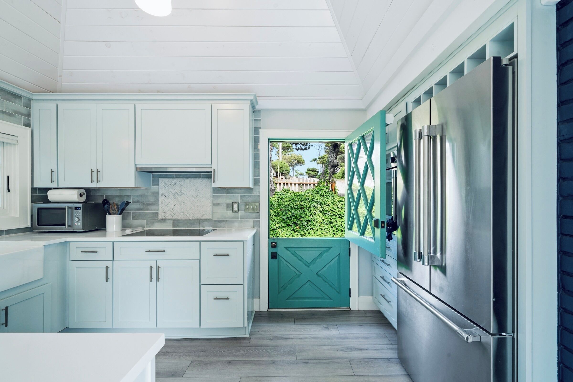 A modern kitchen with light blue cabinets, a stainless steel refrigerator, and a teal door leading to a garden.