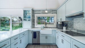 A modern kitchen with light blue cabinets, a stainless steel dishwasher, and a large window overlooking a garden. - Quintessential Renovated Historic Cottage in Neskowin Village + Hot Tub (Neskowin)