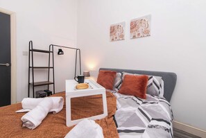 Et pænt opredt soveværelse med en seng, et sengebord med kop og tallerken, et håndklædestativ og to billeder på væggen.