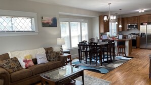 A spacious living room with a brown sofa, a glass coffee table, and a dining area with wooden chairs and a kitchen in the background. - Surfs Up! Pro Cleaned, Self Check in - Sleeps 14 (Racine)