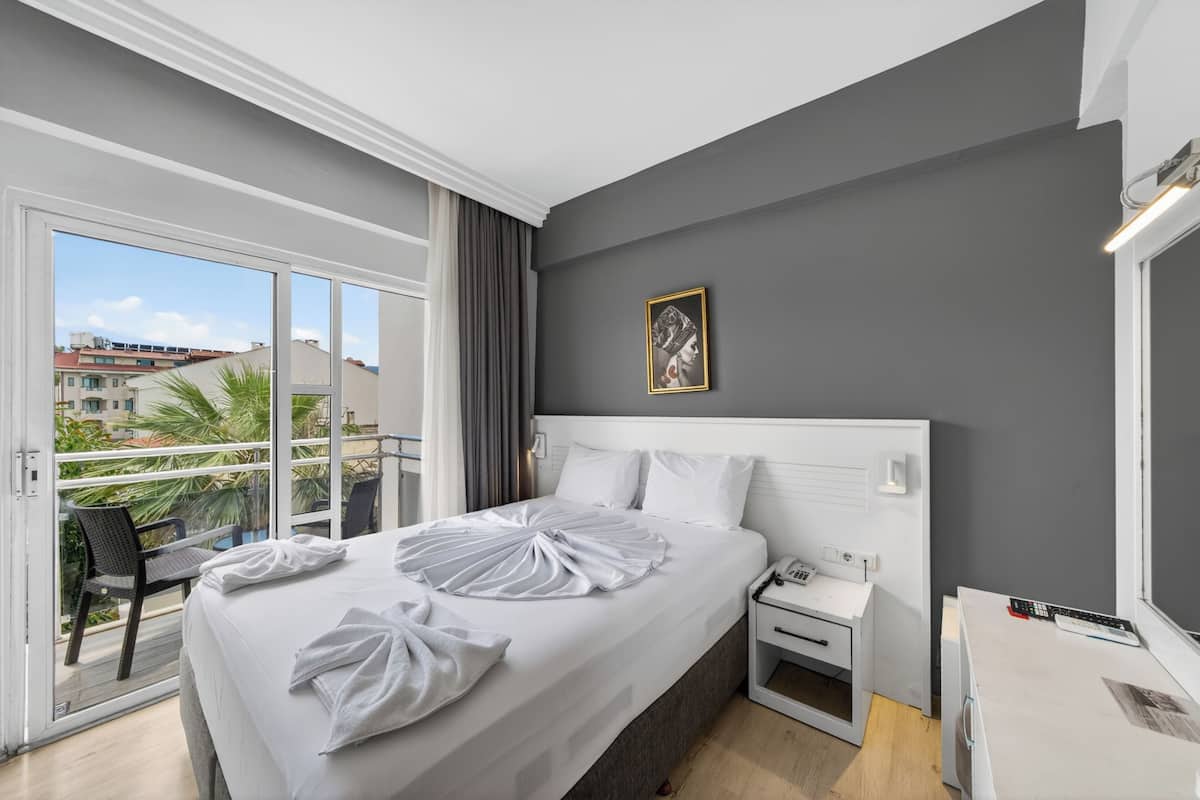 A hotel room with a large bed, a bedside table, a nightstand, a window with a view of buildings, and a balcony with a chair.