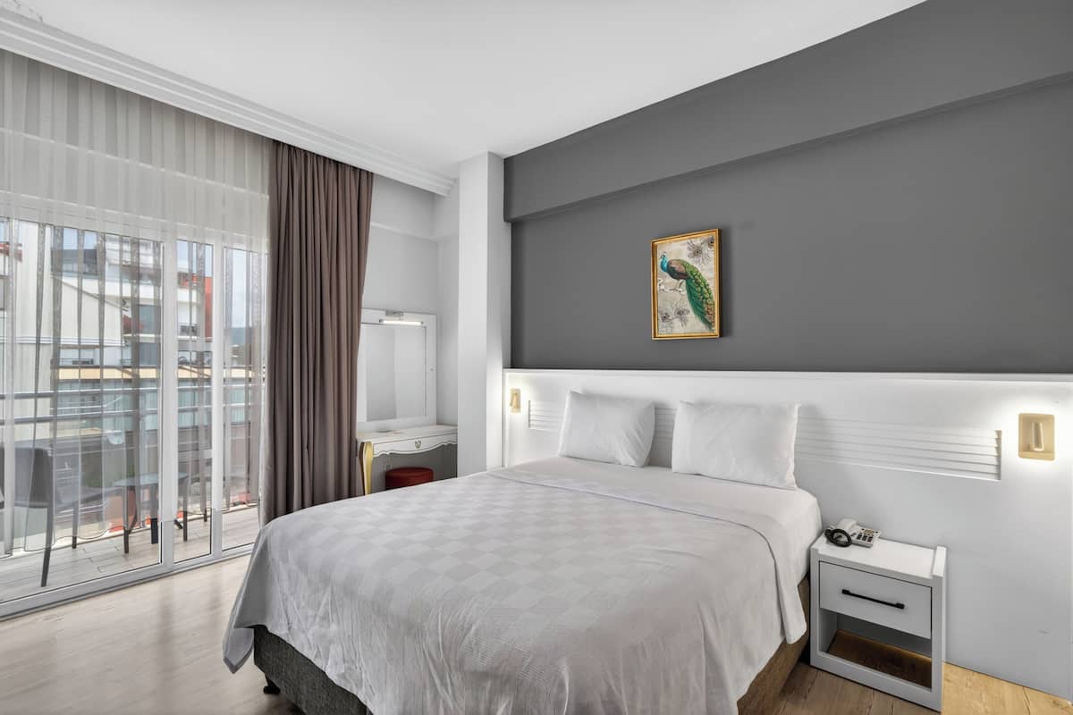 A hotel room with a large bed, a bedside table, a nightstand, a window with curtains, and a painting on the wall.