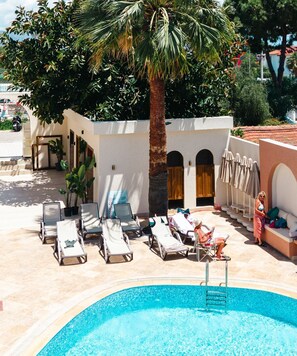 A poolside area with lounge chairs, a palm tree, and a building with arched doorways.