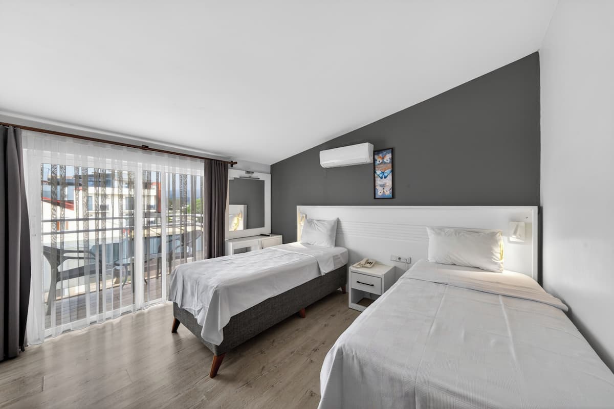 A hotel room with two beds, a balcony with a view, a white air conditioning unit, and a small bedside table with a lamp.