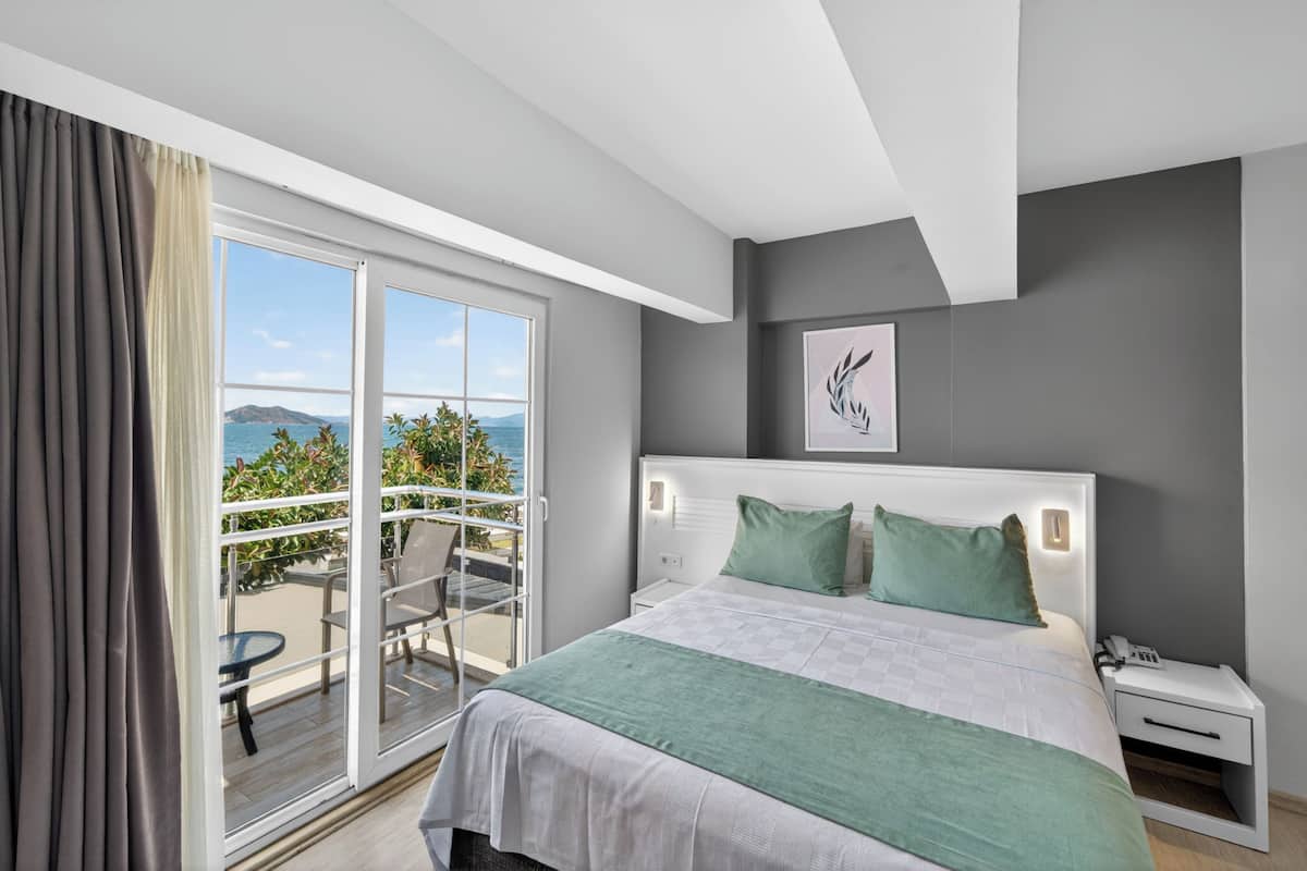 A bedroom with a bed, a nightstand, a phone, a window with a view of the sea, and a balcony.