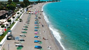 A beach with rows of sun loungers and umbrellas, a promenade with shops, and a view of the sea and mountains in the background. - Mia Casa Hotel (Fethiye)