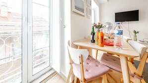 Une pièce lumineuse avec une table en bois, deux chaises, un téléviseur fixé au mur et un balcon offrant une vue sur les immebles.