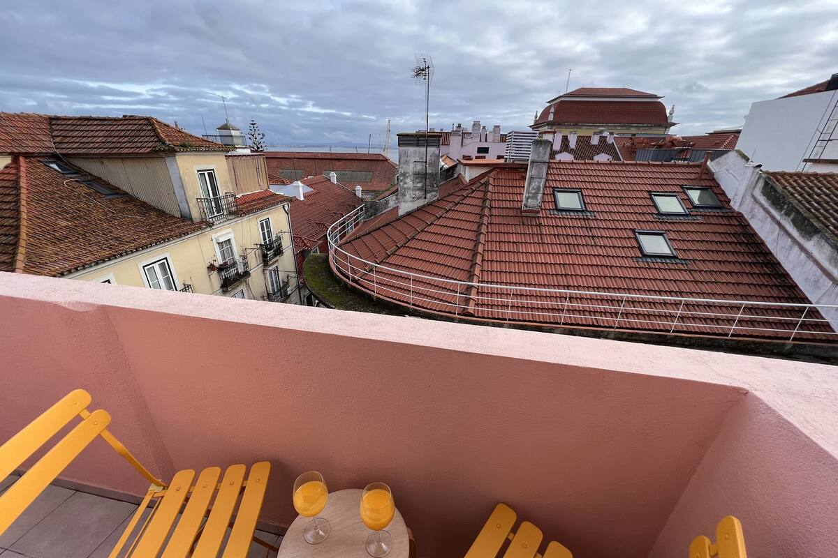 Un balcon avec des chaises jaunes et une petite table, donnant sur un paysage urbain avec des toits recouverts de tuiles rouges et un ciel nuageux.