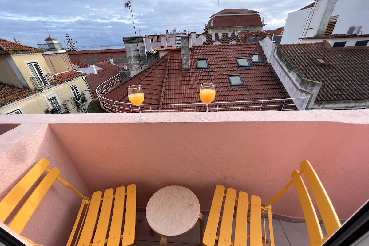 Un balcon avec des chaises jaunes et une petite table ronde, donnant sur un paysage urbain avec des toits recouverts de tuiles rouges et une vue sur la mer.