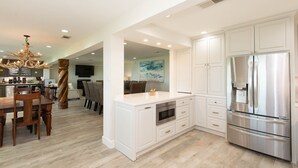 A modern kitchen with white cabinetry, a stainless steel refrigerator, and a dining area with wooden chairs and a chandelier. - Stunning Five Bedroom Escape - Great Water! (Enchanted Oaks)