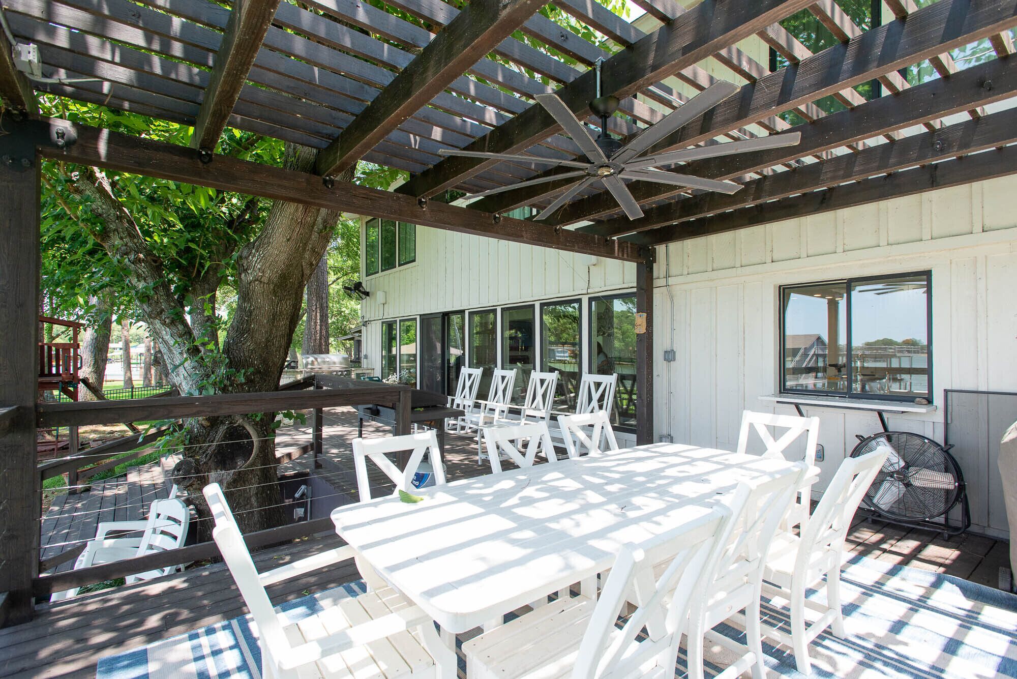 An outdoor patio with white tables and chairs, a ceiling fan, and a view of a building in the background.