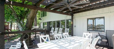 An outdoor patio with white tables and chairs, a ceiling fan, and a view of a building in the background.