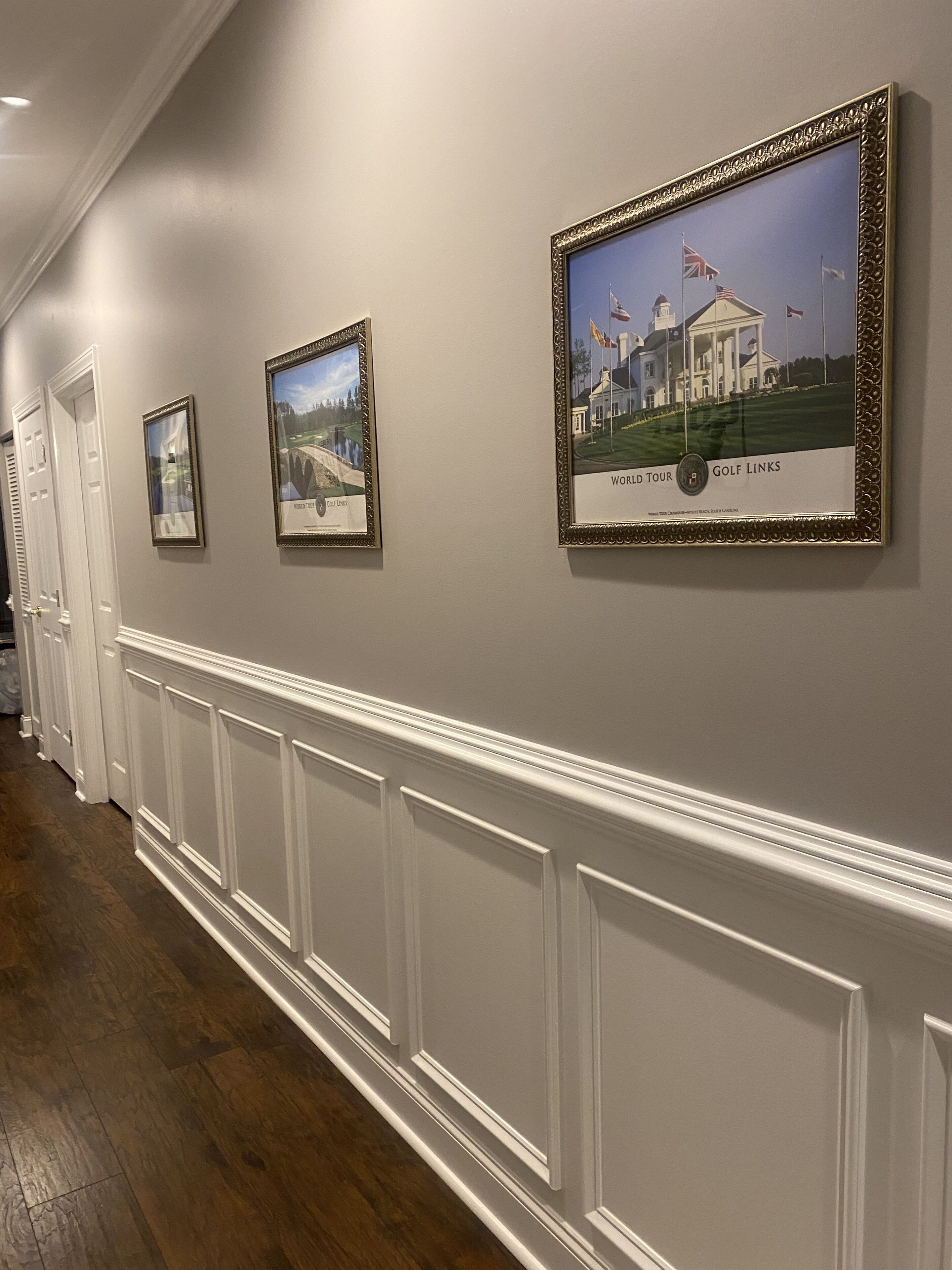 Un couloir avec des tableaux encadrés accrochés au mur, des lambris blancs et un plancher en bois.