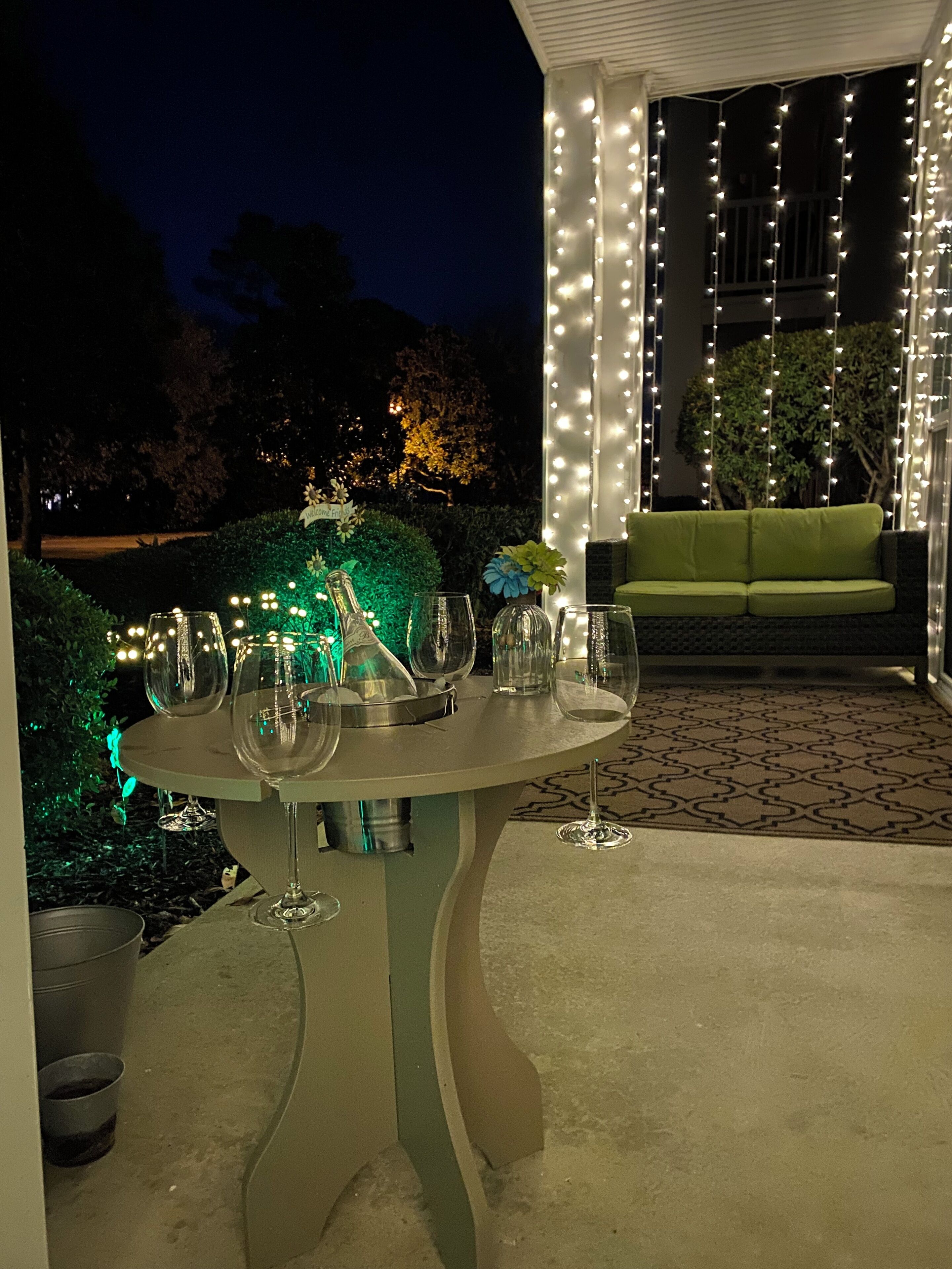An outdoor patio with a round table set for dining, surrounded by string lights and greenery.