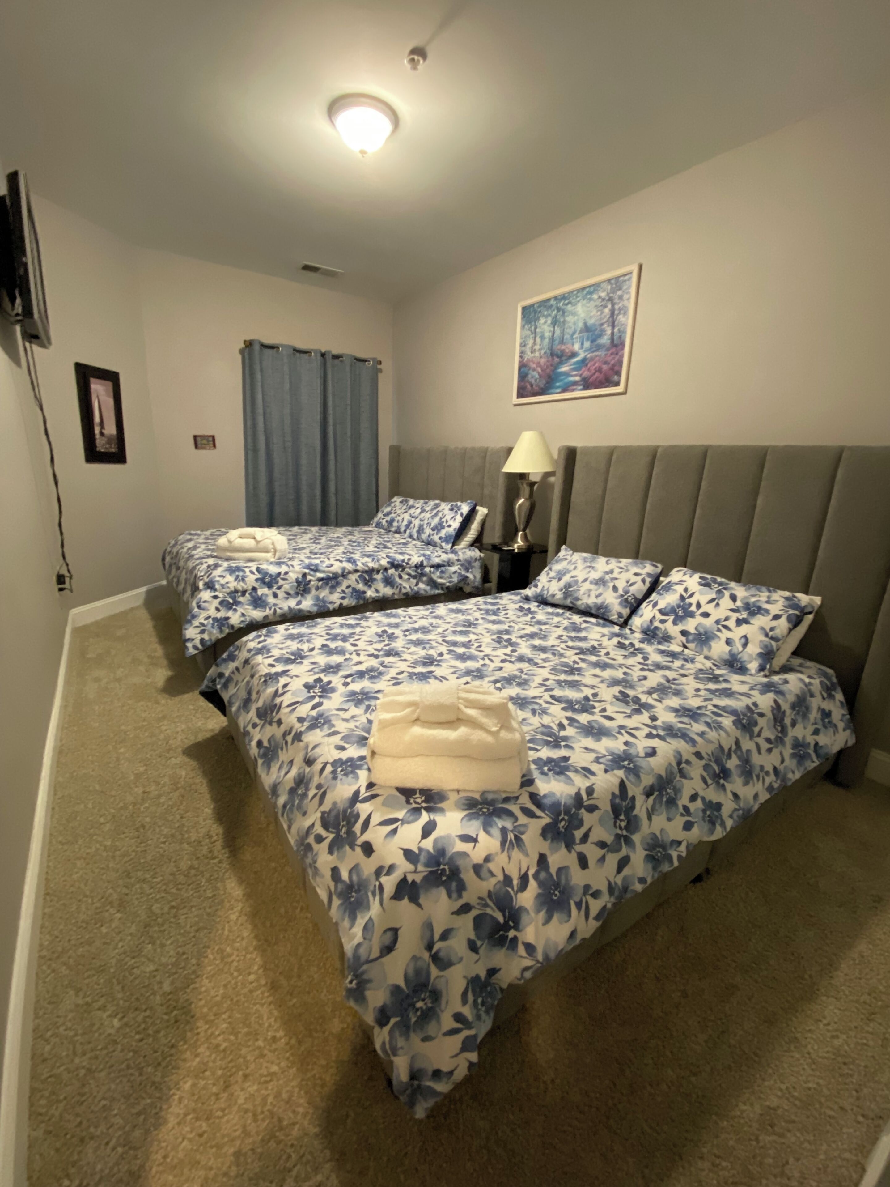 A hotel room with two beds, a nightstand, a lamp, a painting, and a ceiling light.