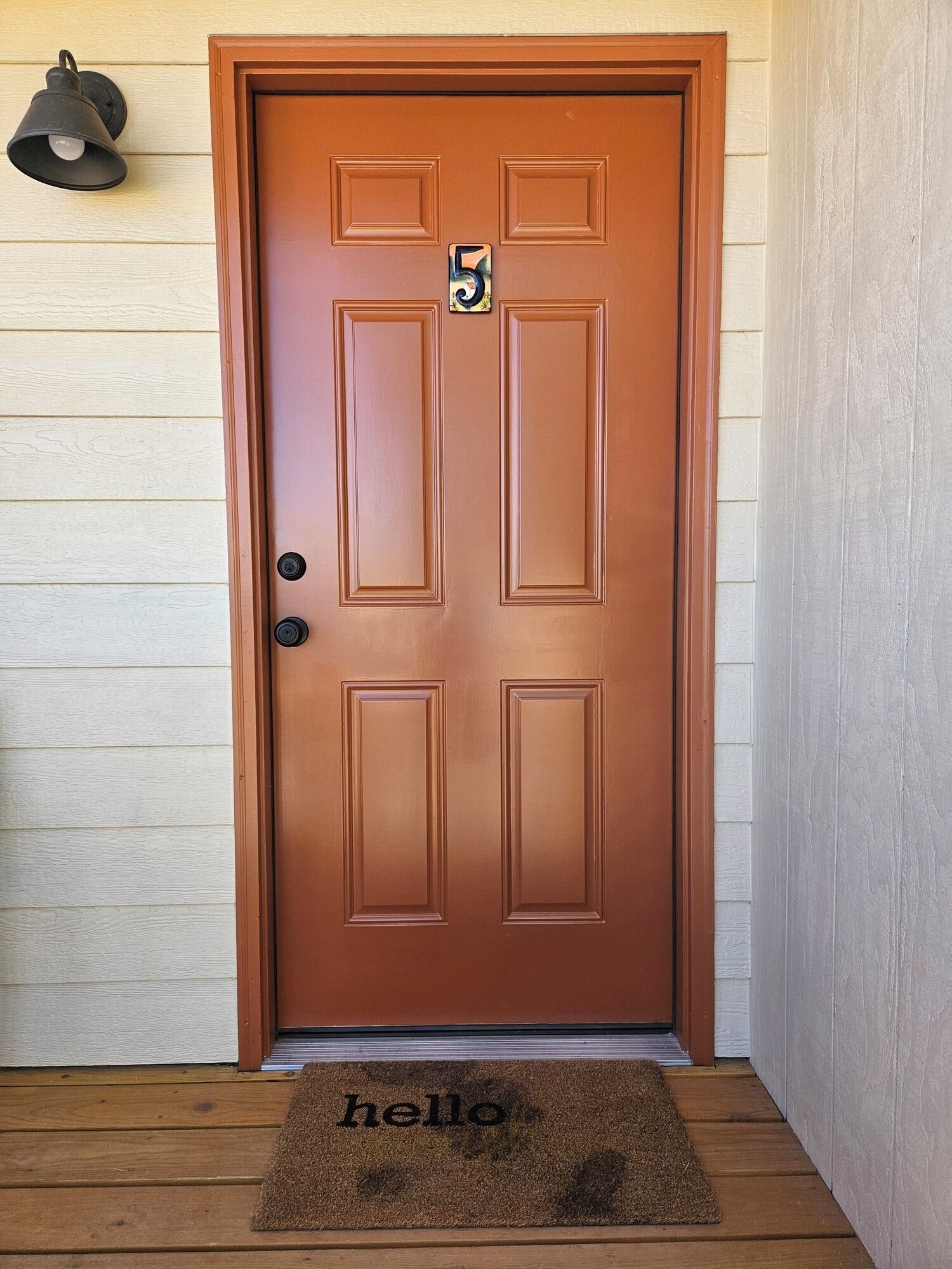 Una puerta marrón con un cartel con el número 5, un felpudo que dice 'hello' y una lámpara de pared.
