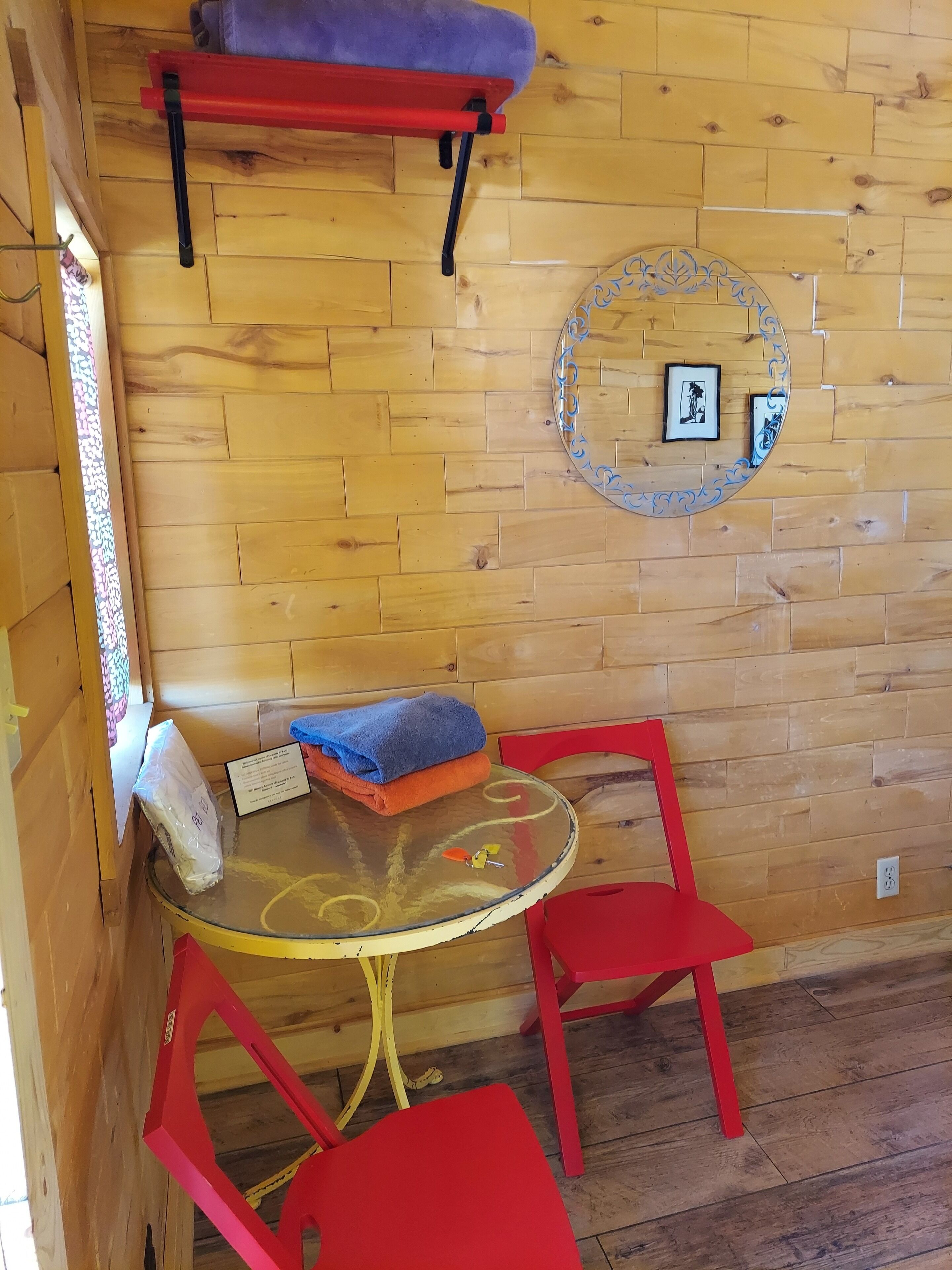 A wooden cabin interior with a round glass table, red chairs, a red shelf, and a decorative wall plate.