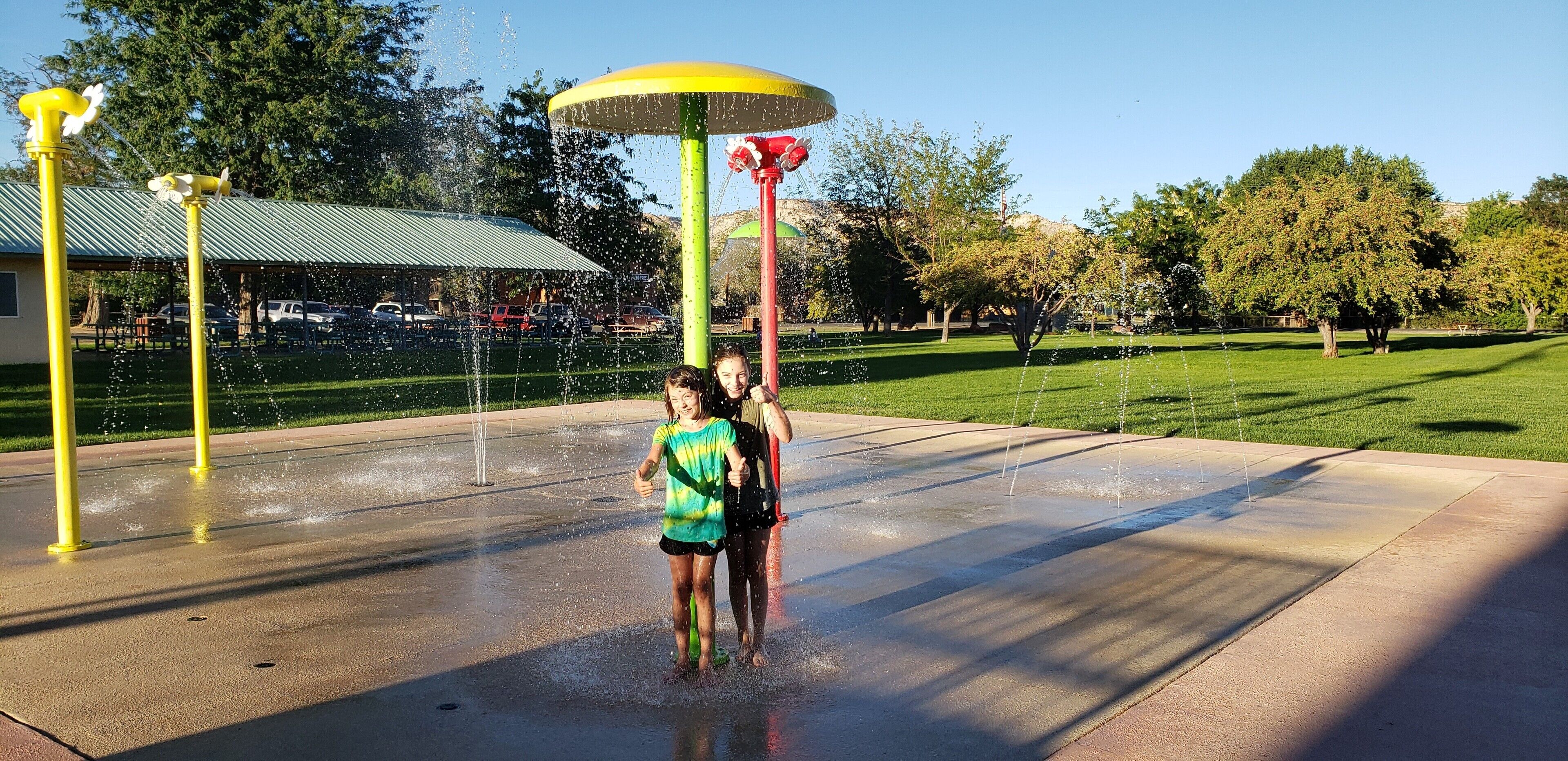 A water feature with multiple jets, a green umbrella, and a red structure in a park setting.