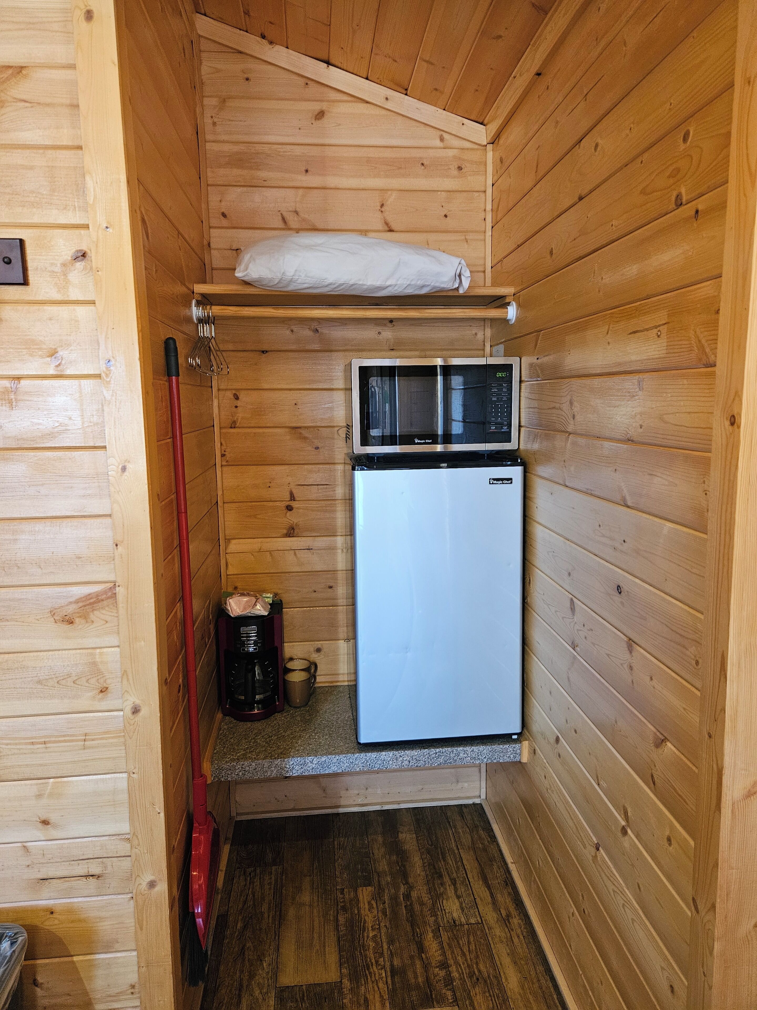 Une cuisine compacte équipée d’un four à micro-ondes et d’un petit réfrigérateur, avec des murs en bois et un balai rouge appuyé contre le mur.