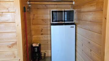 Une cuisine compacte équipée d’un four à micro-ondes et d’un petit réfrigérateur, avec des murs en bois et un balai rouge appuyé contre le mur.