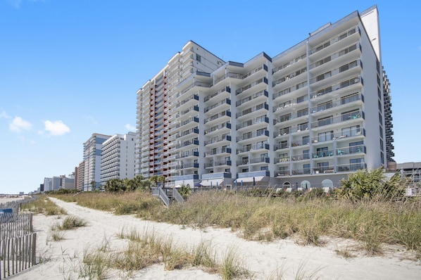 A high-rise hotel with multiple balconies, a sandy beach in the foreground, and a clear blue sky.