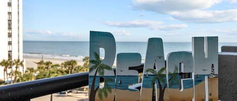 A wooden sign with the word "REACH" and beach-themed decorations, overlooking a beach with palm trees and a lighthouse.
