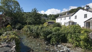Un bâtiment blanc surmonté d’un toit en pierre, entouré de verdure et bordé par un ruisseau aux eaux claires.