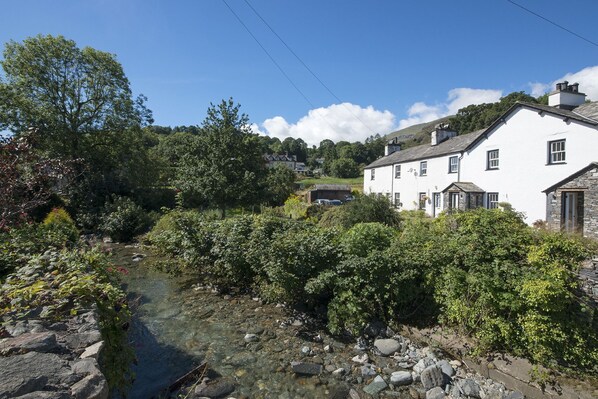 A white building with a stone roof, surrounded by greenery and a clear stream. - No. 3 The Forge, village cottage in quiet location with walks from the door (Coniston)