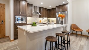 Une cuisine moderne avec des meubles en bois, un réfrigérateur en acier inoxydable, un plan de travail blanc et deux tabourets de bar.