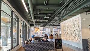 Un intérieur doté de grandes fenêtres, d’un haut plafond avec des tuyaux apparents, d’un espace salon avec des tables et d’un mur décoré d’assiettes.