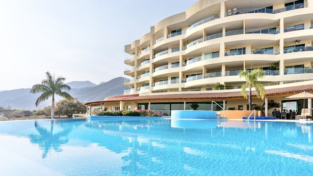 Un hotel de varios pisos con piscina, balcones y montañas al fondo.