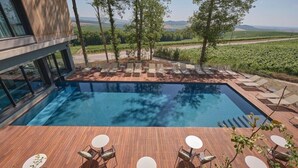 Zona de piscina moderna con sillones reclinables y mesas, rodeada de una terraza de madera y con vistas a un viñedo.