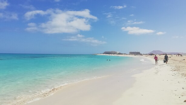 A beach with clear turquoise water, white sand, and a few people walking along the shore.