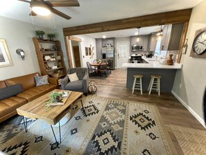 A spacious living room with a brown leather sofa, a wooden coffee table, and a patterned rug. A kitchen with a bar area is visible in the background.