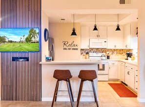A modern kitchen with white cabinets, a built-in oven, and a red rug. There are two bar stools and a TV mounted on the wall.