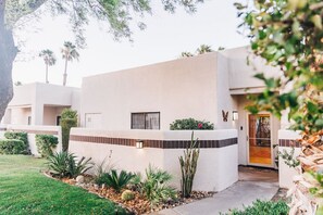 Un edificio moderno con techo plano, una entrada pequeña y un jardín bien cuidado con diversas plantas y un cactus.